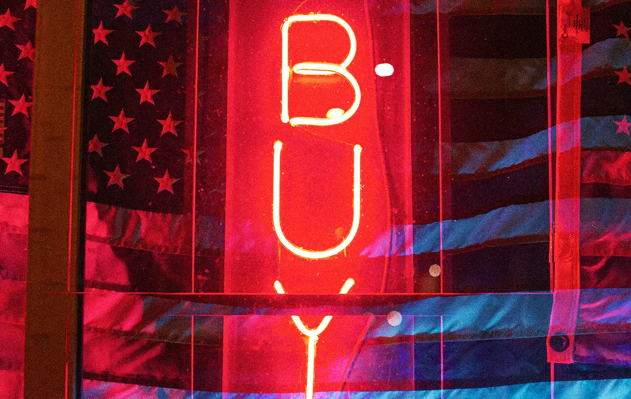 Bold red neon “BUY” sign layered over an American flag, symbolizing Buy American initiatives, domestic manufacturing, and U.S.-made product sourcing.