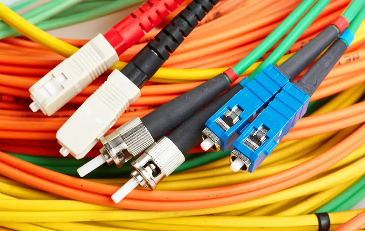 Close-up of colorful fiber optic cables (orange, yellow, and green) with multiple connector types, including blue, white, and metal-tipped ends, bundled together in a network wiring setup.