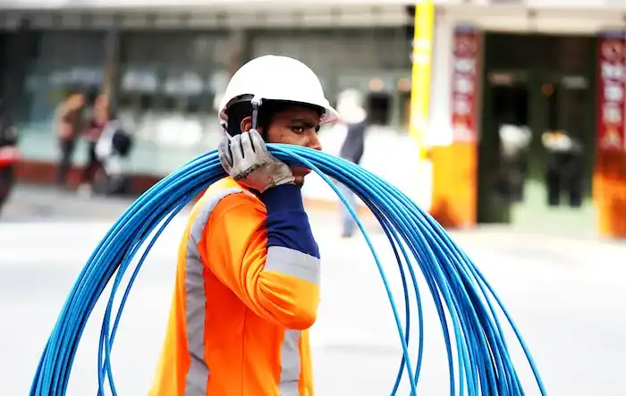 Construction worker carrying blue cables wearing safety gear on a city street.