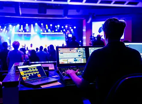 Live event production technician managing audio, video, and lighting systems from a control booth during a stage performance.