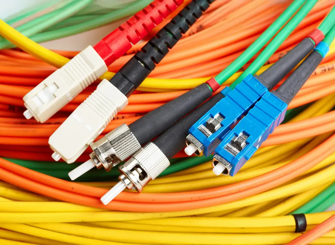 Close-up of colorful fiber optic cables (orange, yellow, and green) with multiple connector types, including blue, white, and metal-tipped ends, bundled together in a network wiring setup.